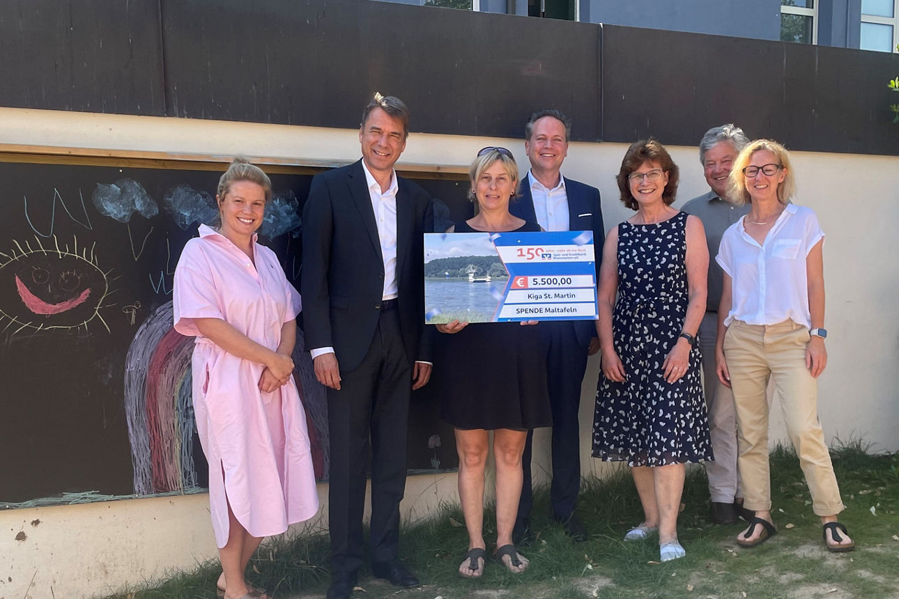 Spendenübergabe, sieben Personen vor einem Gebäude mit einem Scheck. 