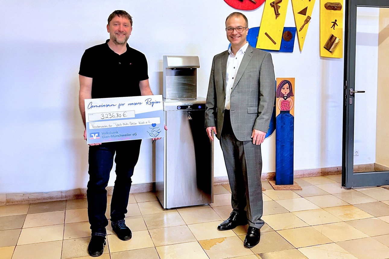 Zwei Männer vor einem Wasserspender. Der Mann rechts trägt einen grauen Anzug. Der Mann rechts hält einen Geldscheck fest.