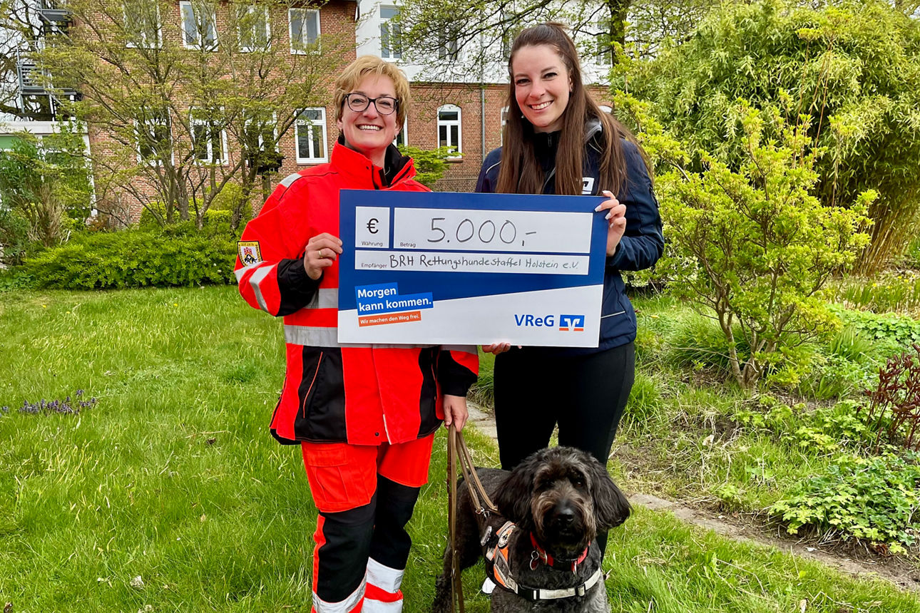 Zwei Frauen halten lächelnd einen Spendencheck. Vor ihnen steht ein Hund.