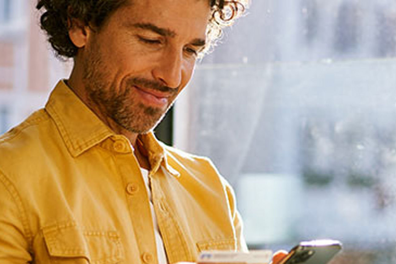 Ein Mann mit lockigem Haar und gelbem Hemd hält in einer Hand ein Smartphone und in der anderen eine Bankkarte.