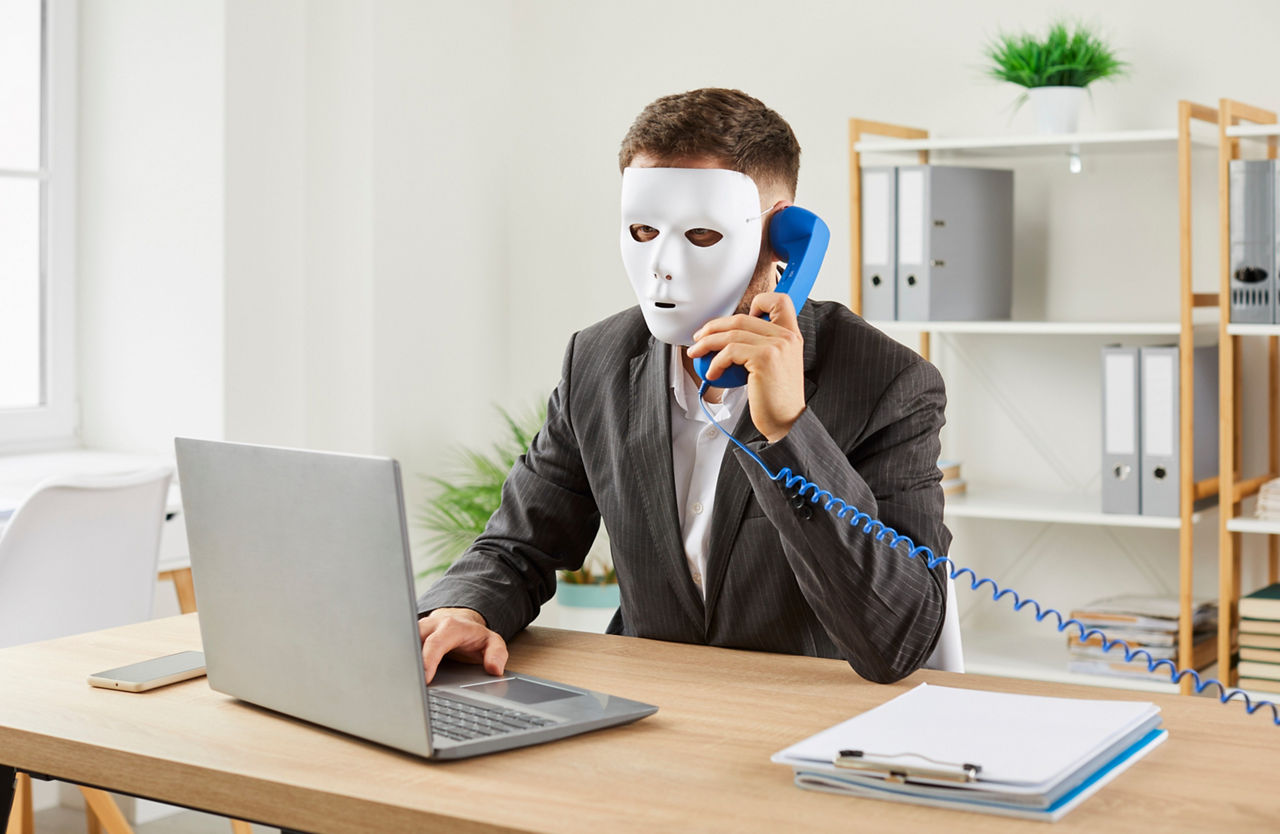 Betrüger mit Maske am Telefon hinter Computer