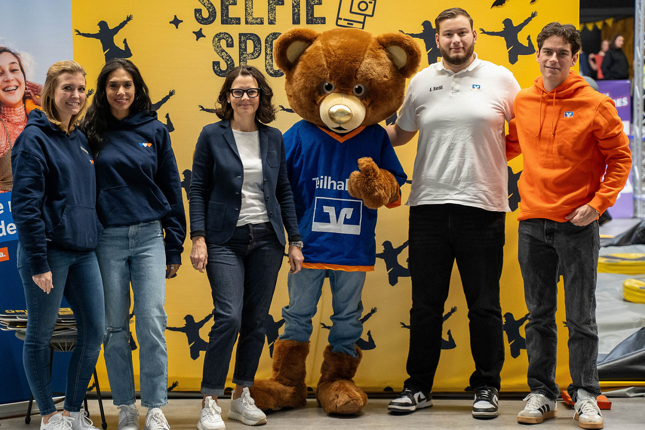 Gruppe vor gelbem Selfie-Spot-Hintergrund posiert mit TeilhaBÄR in der Mitte.