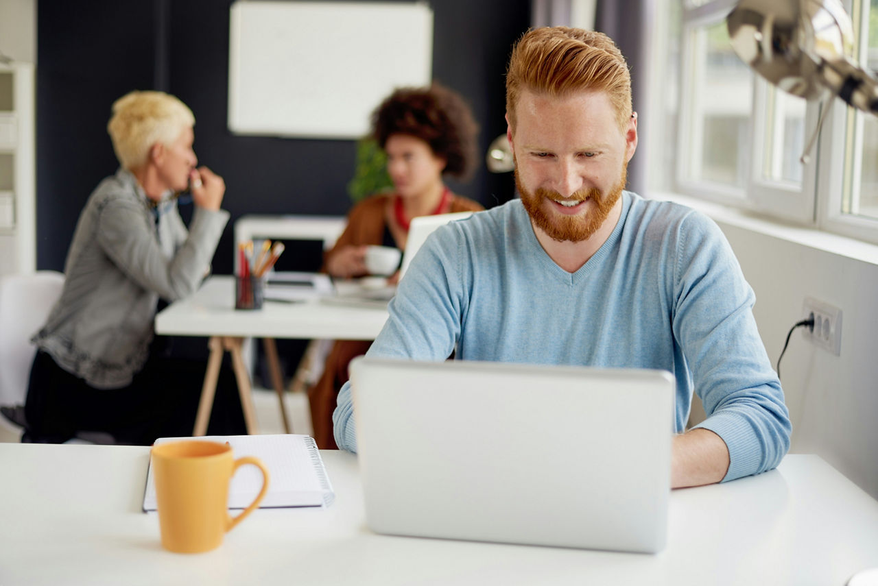 Ein Mann sitzt allein mit Laptop am Tisch, zwei Frauen unterhalten sich im Hintergrund