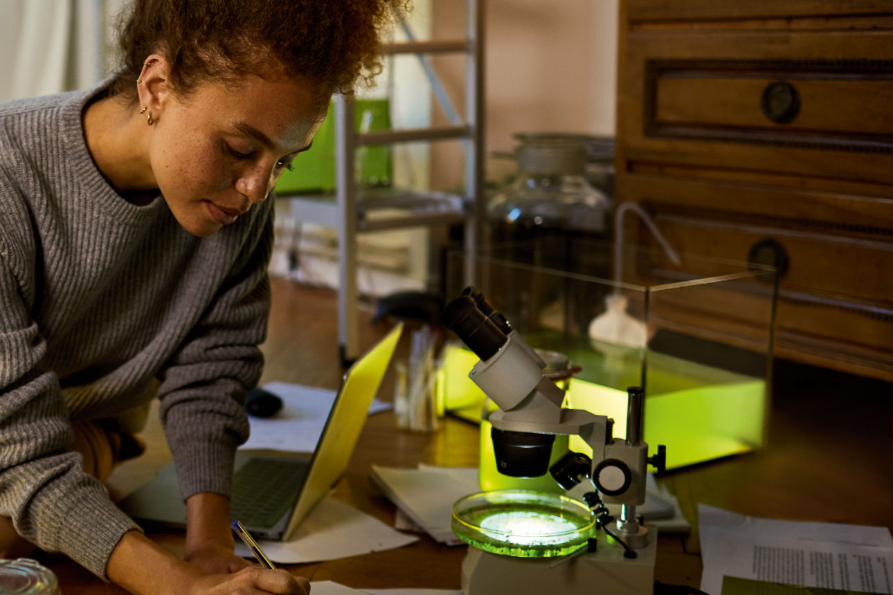 Junge Frau sitzt mit dem Laptop auf dem Boden und schreibt mit einem Stift in ein Buch.