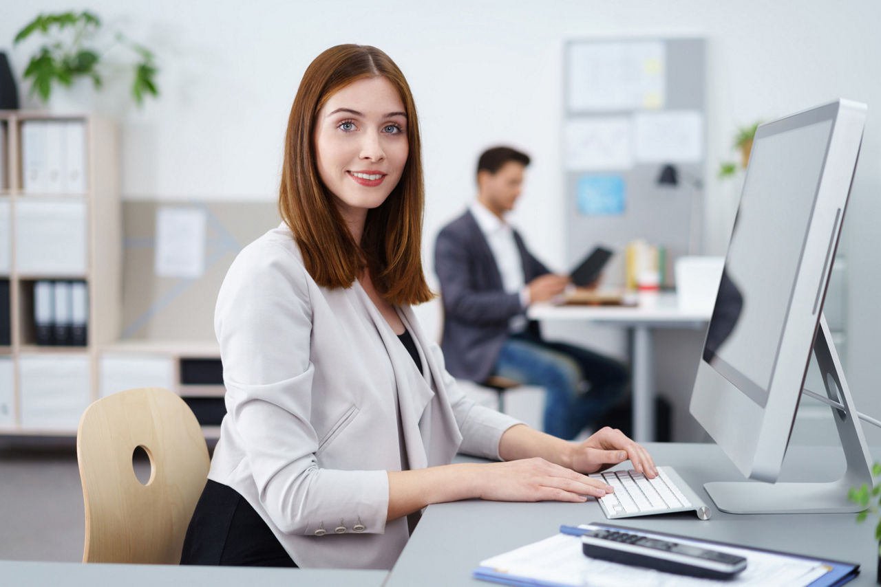 Frau sitzt im Büro am PC und lächelt in die Kamera
