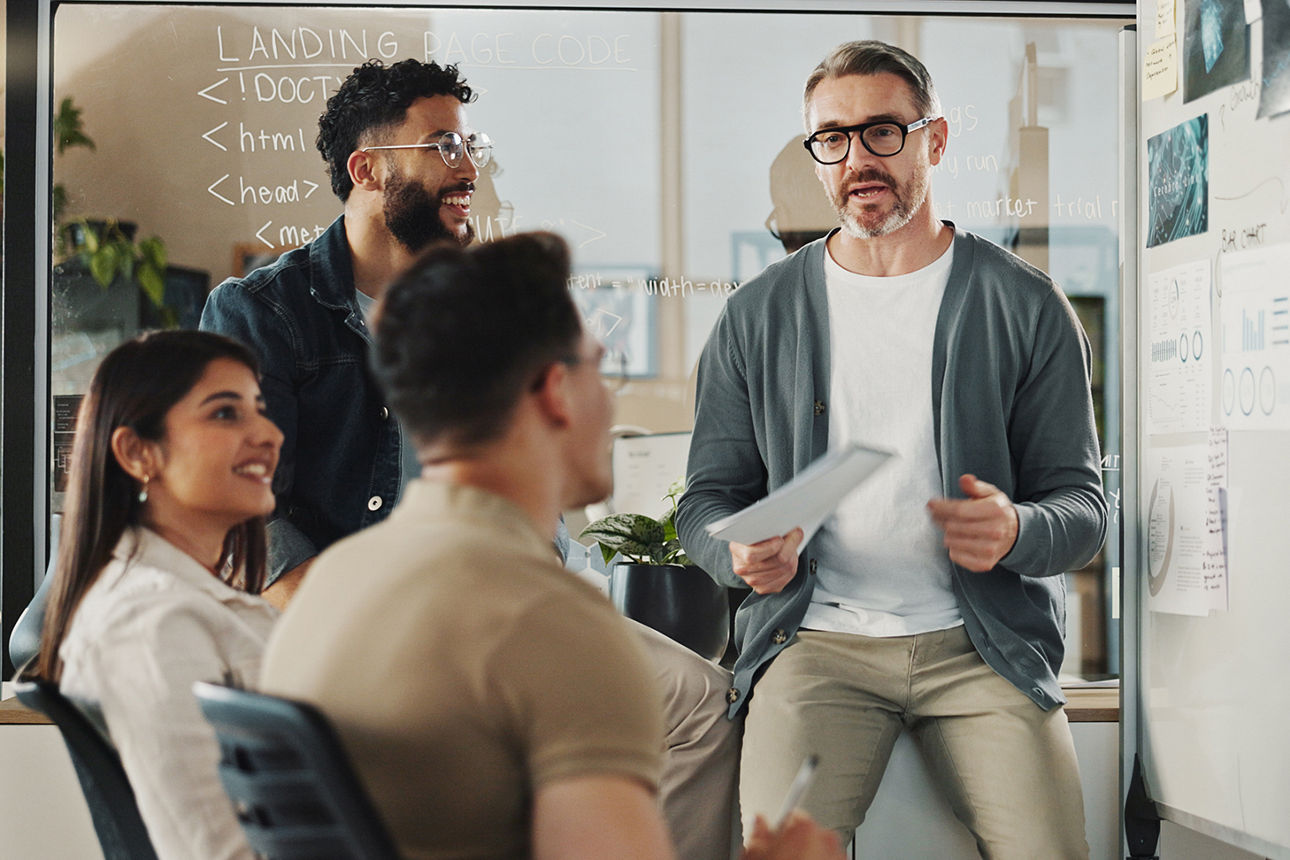 Ein Team vor einer Glaswand an einem Whiteboard. Ein Mann und eine Frau sitzen. Zwei Männer stehen.
