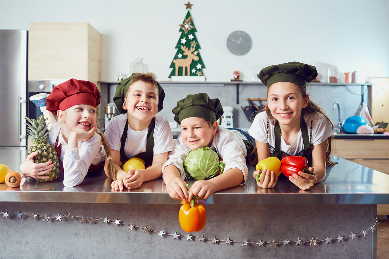 Vier fröhliche Kinder mit Kochmützen in einer Küche, sie halten verschiedene Sorten Obst und Gemüse in den Händen.