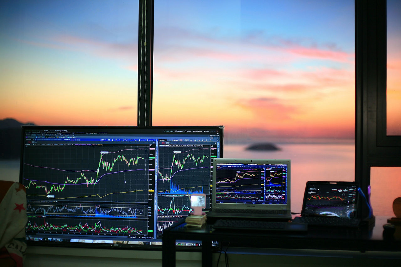 Laptops mit Trading Charts stehen vor einem Fenster, im Hintergrund sieht man das Meer und den Sonnenuntergang