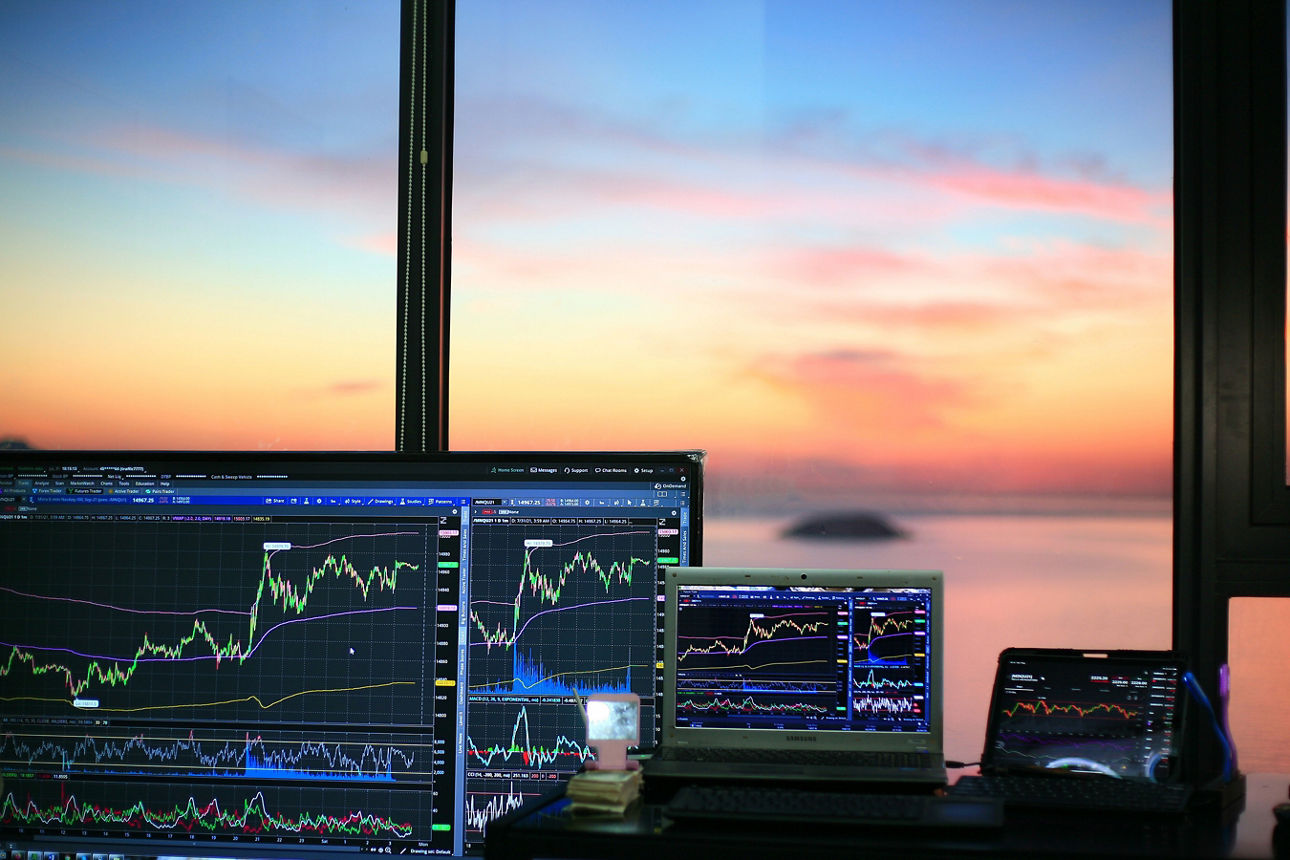 Laptops mit Trading Charts stehen vor einem Fenster, im Hintergrund sieht man das Meer und den Sonnenuntergang