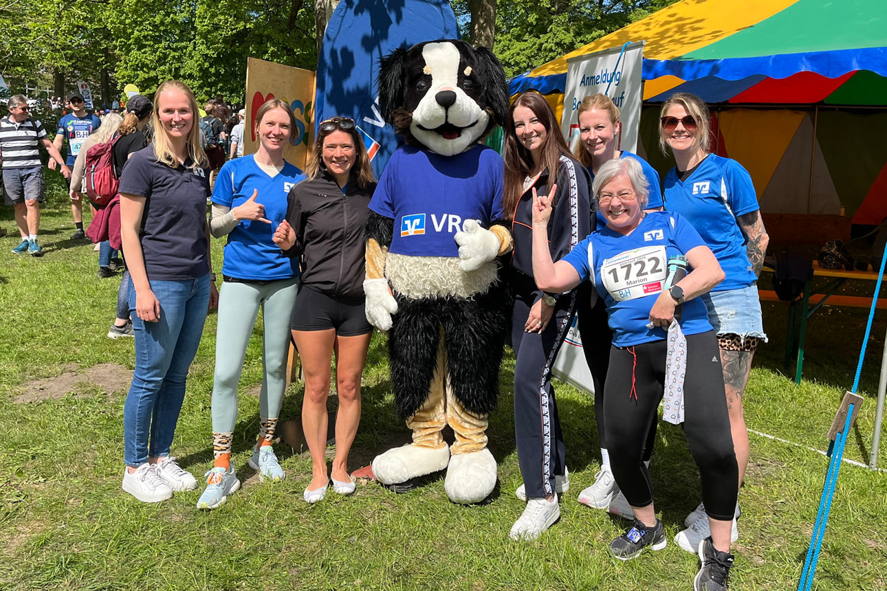 Viele VReG Mitarbeitende im sportlichen Outfit stehen als Gruppe mit dem Maskottchen "Bankhund Anton" auf einer Wiese.