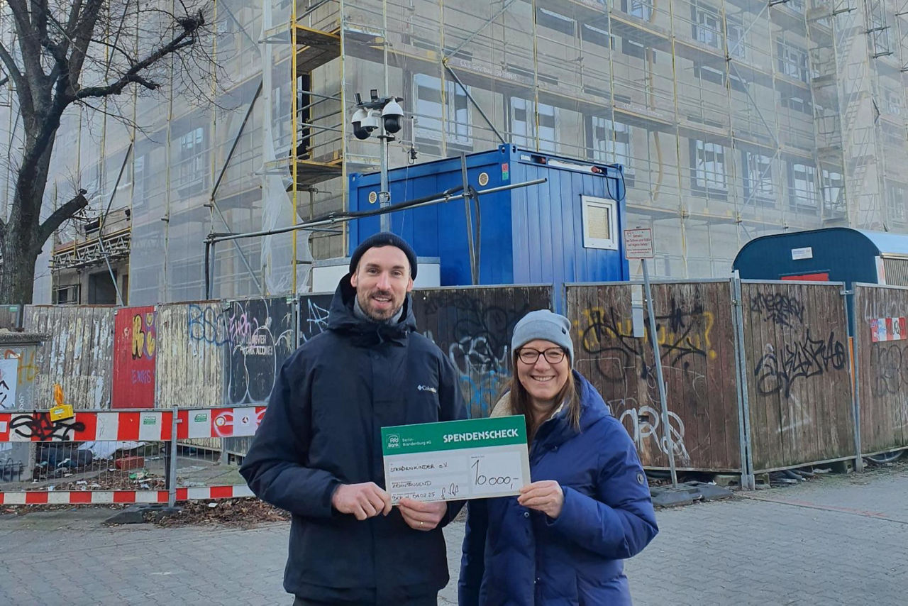 Mann und Frau mit Spendenscheck in den Händen stehen vor Baustelle eines Neubaus