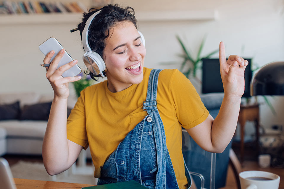 Eine junge Frau sitzt gestikulierend und freudestrahlend im Wohnzimmer und hört Musik mit ihren Kopfhörern