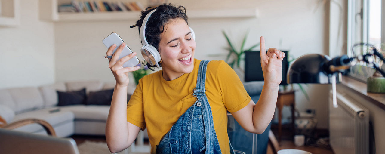 Eine junge Frau sitzt gestikulierend und freudestrahlend im Wohnzimmer und hört Musik mit ihren Kopfhörern
