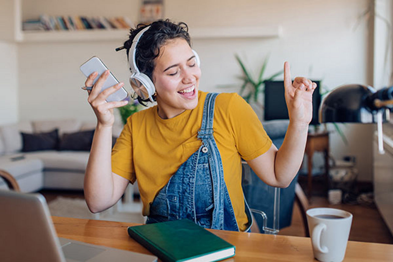 Eine junge Frau sitzt gestikulierend und freudestrahlend im Wohnzimmer und hört Musik mit ihren Kopfhörern