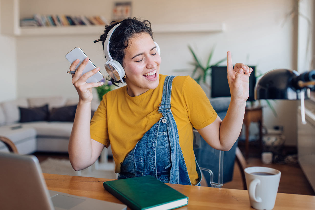Eine junge Frau sitzt gestikulierend und freudestrahlend im Wohnzimmer und hört Musik mit ihren Kopfhörern