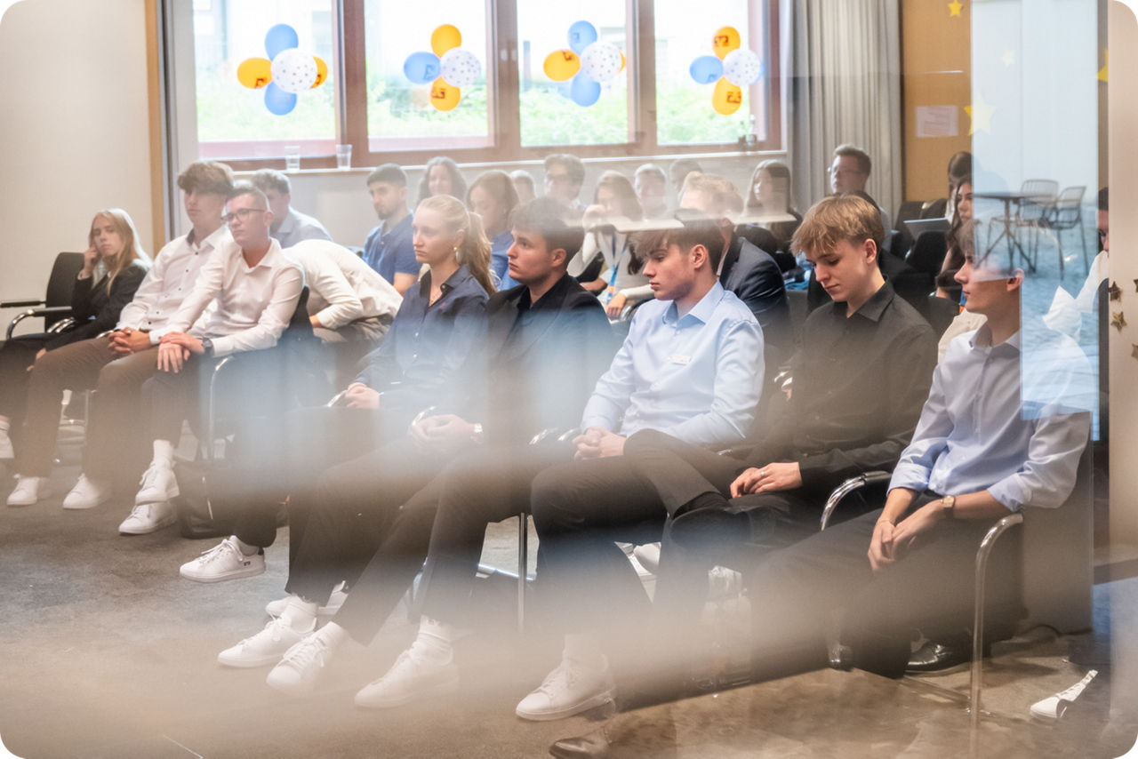 Jugendliche sitzen in einem Vortrag und werden durch eine Scheibe fotografiert