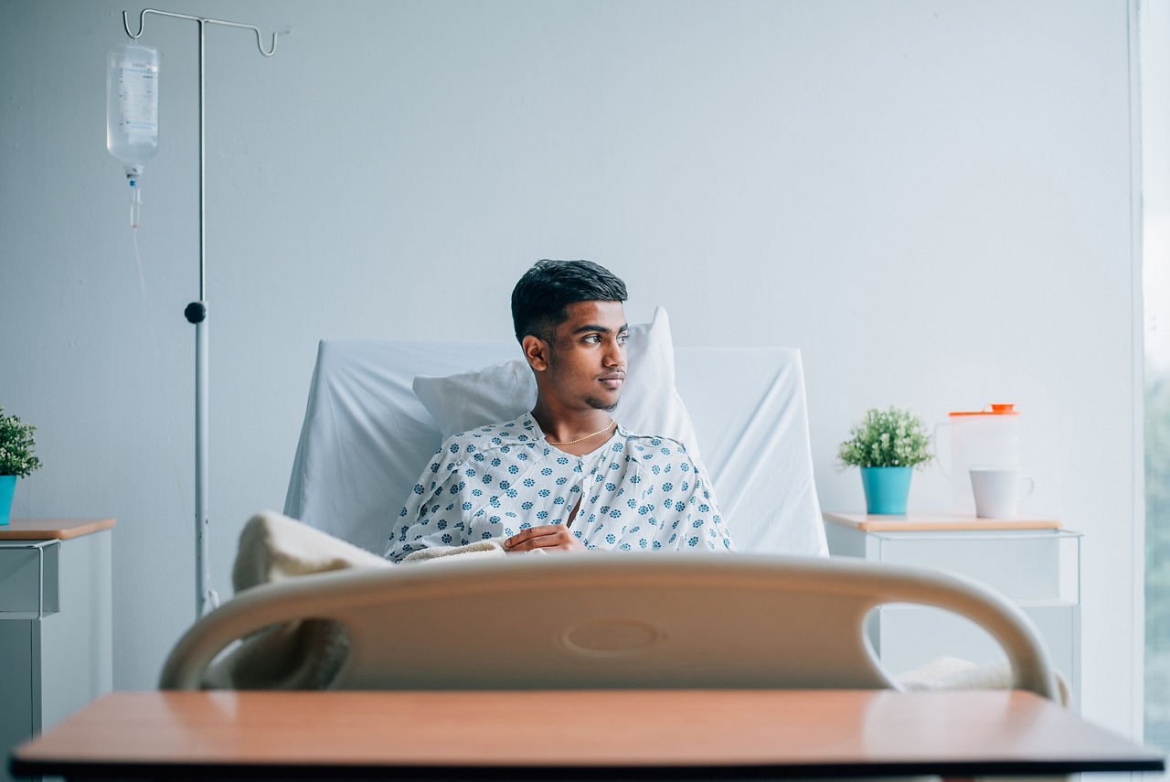 Ein junger Mann sitzt in einem hellen Krankenzimmer aufrecht im Bett. Er blickt in Richtung Fenster. 