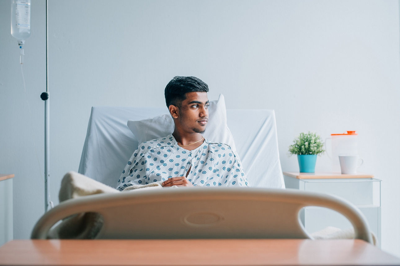 Ein junger Mann sitzt in einem hellen Krankenzimmer aufrecht im Bett. Er blickt in Richtung Fenster. 