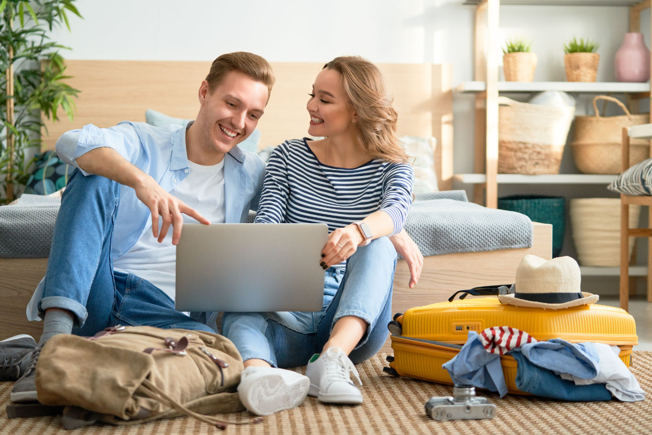 Eine Frau und ein Mann sitzen zwischen Koffern auf dem Boden und schauen auf einen Laptop.