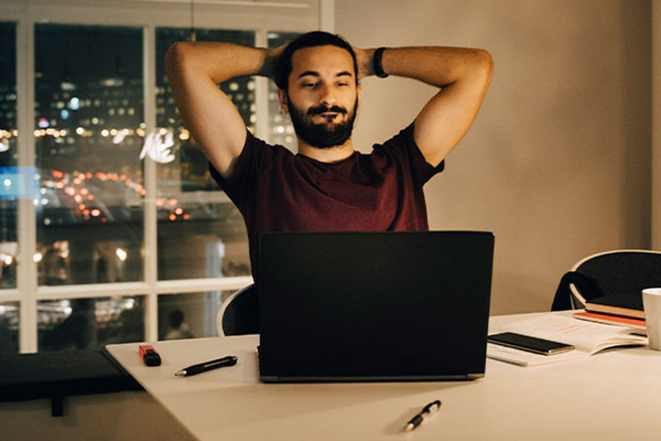 Mann an einem Schreibtisch schaut auf seinen Laptop und lehnt sich entspannt zurück