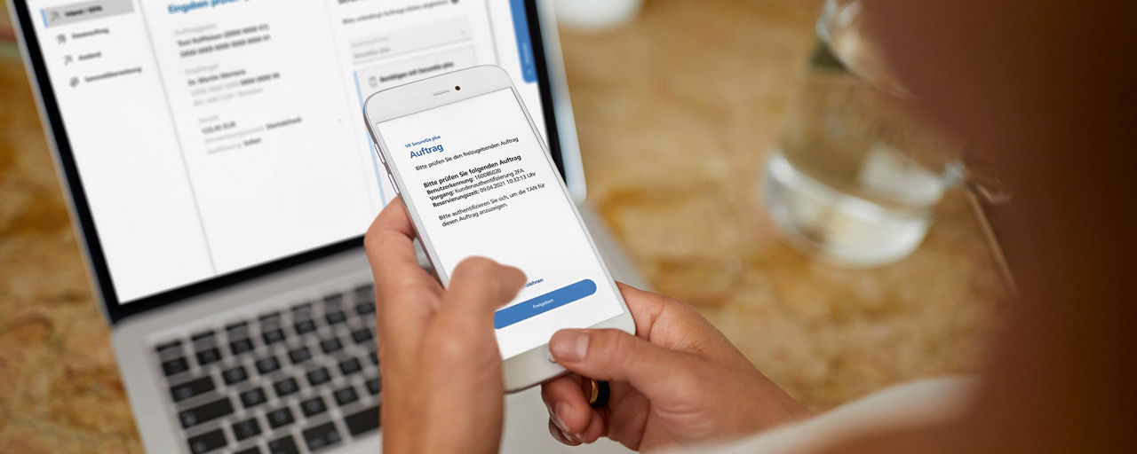 Zu sehen sind ein Laptop und ein Handy welches in der Hand gehalten wird, auf dem ein TAN-Verfahren angewand wird.