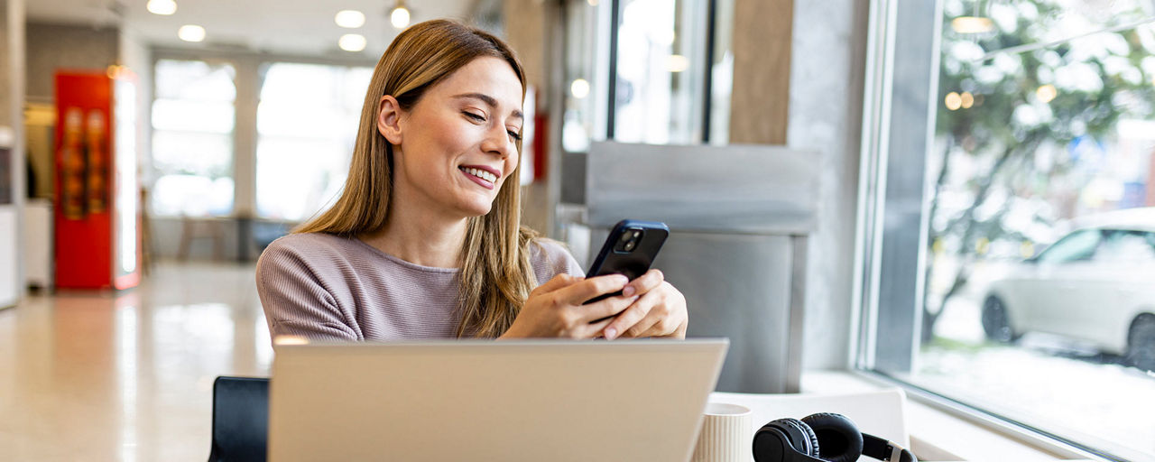 Junge Frau vor einem Laptop, mit einem Smartphone in der Hand