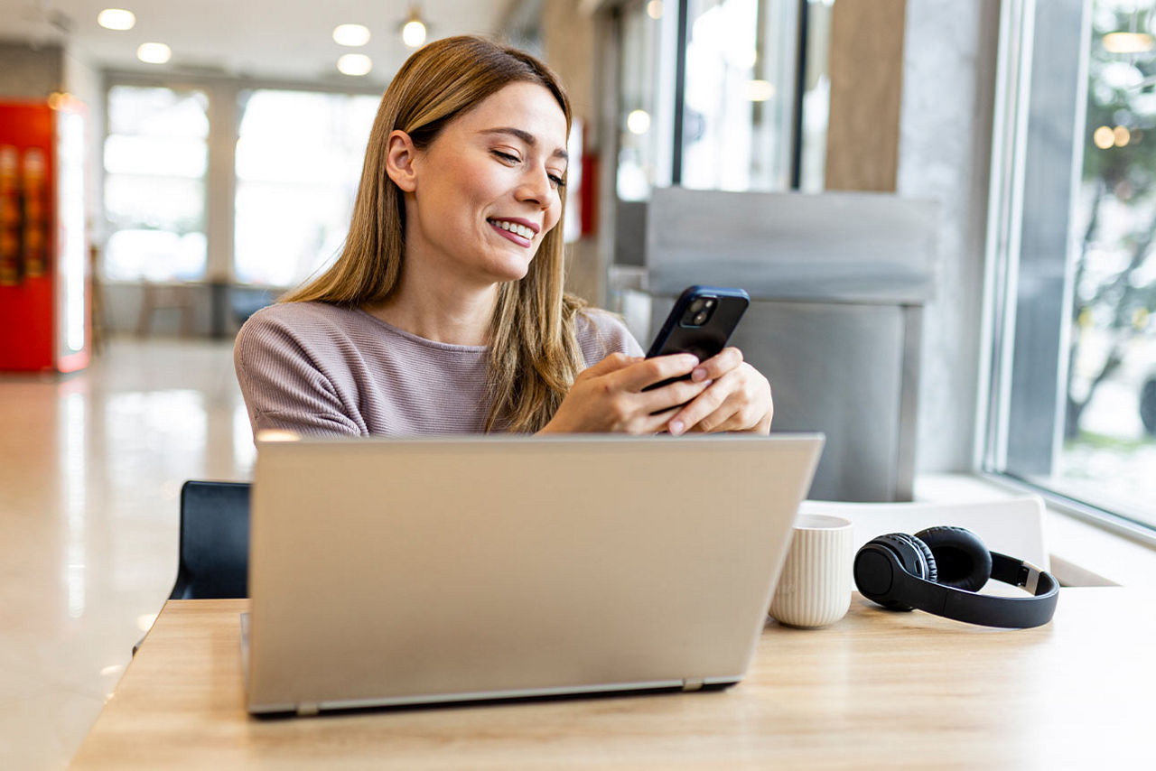 Junge Frau vor einem Laptop, mit einem Smartphone in der Hand