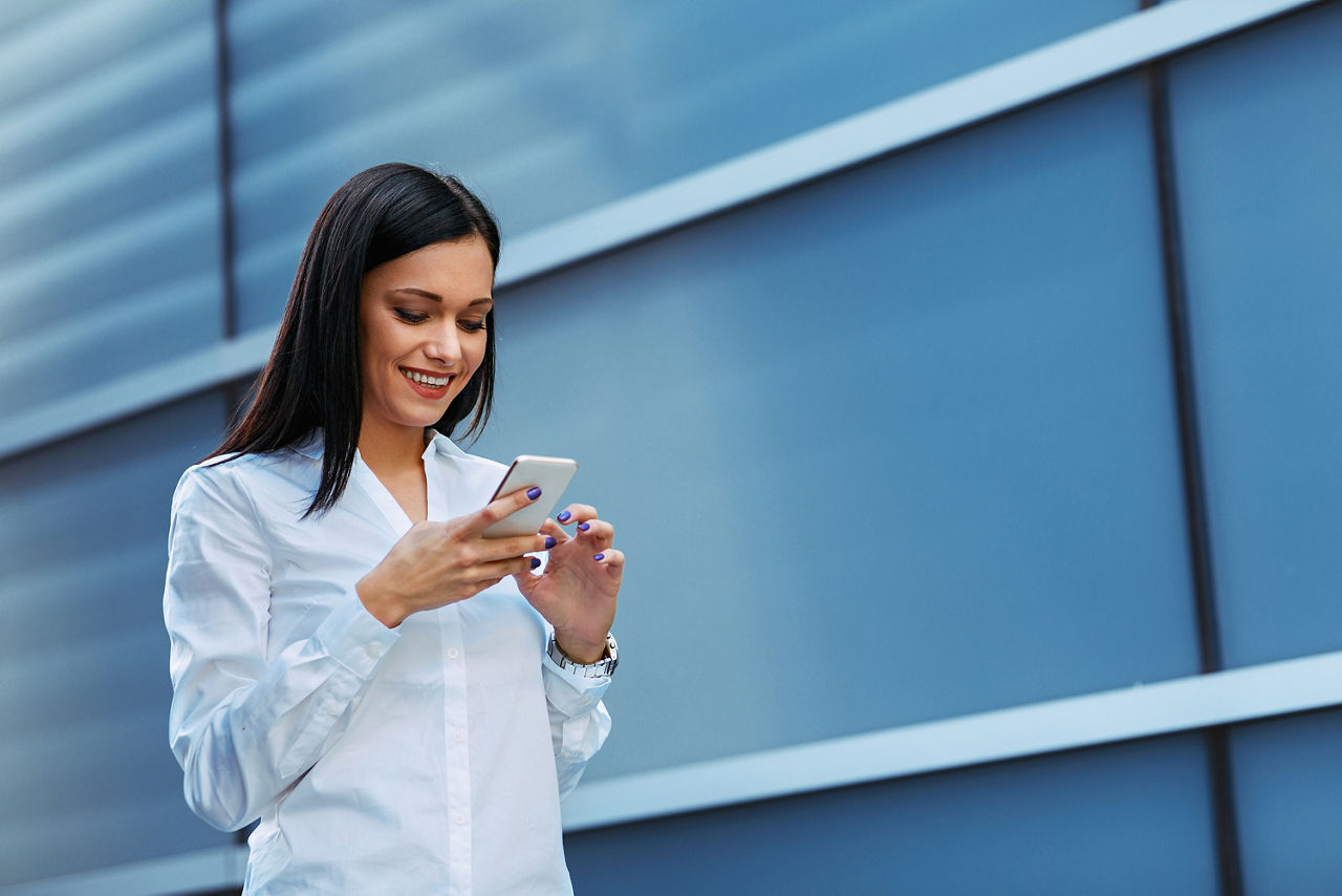 Eine junge Geschäftsfrau vor einem blauen Gebäude schaut lächelnd in ihr Handy