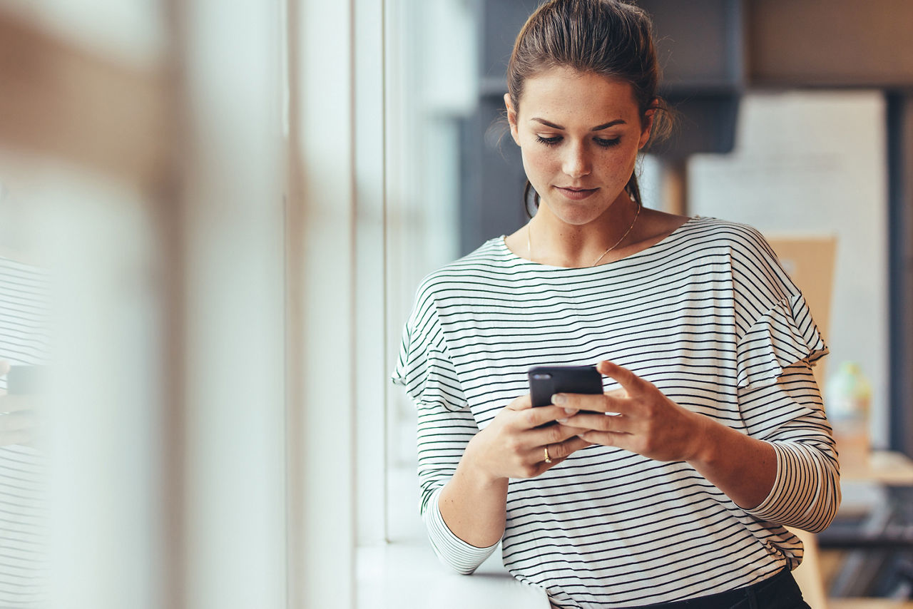 Eine junge Frau mit einem gestreiften Oberteil lehnt sich an der Fensterbank und schaut auf ihr Handy
