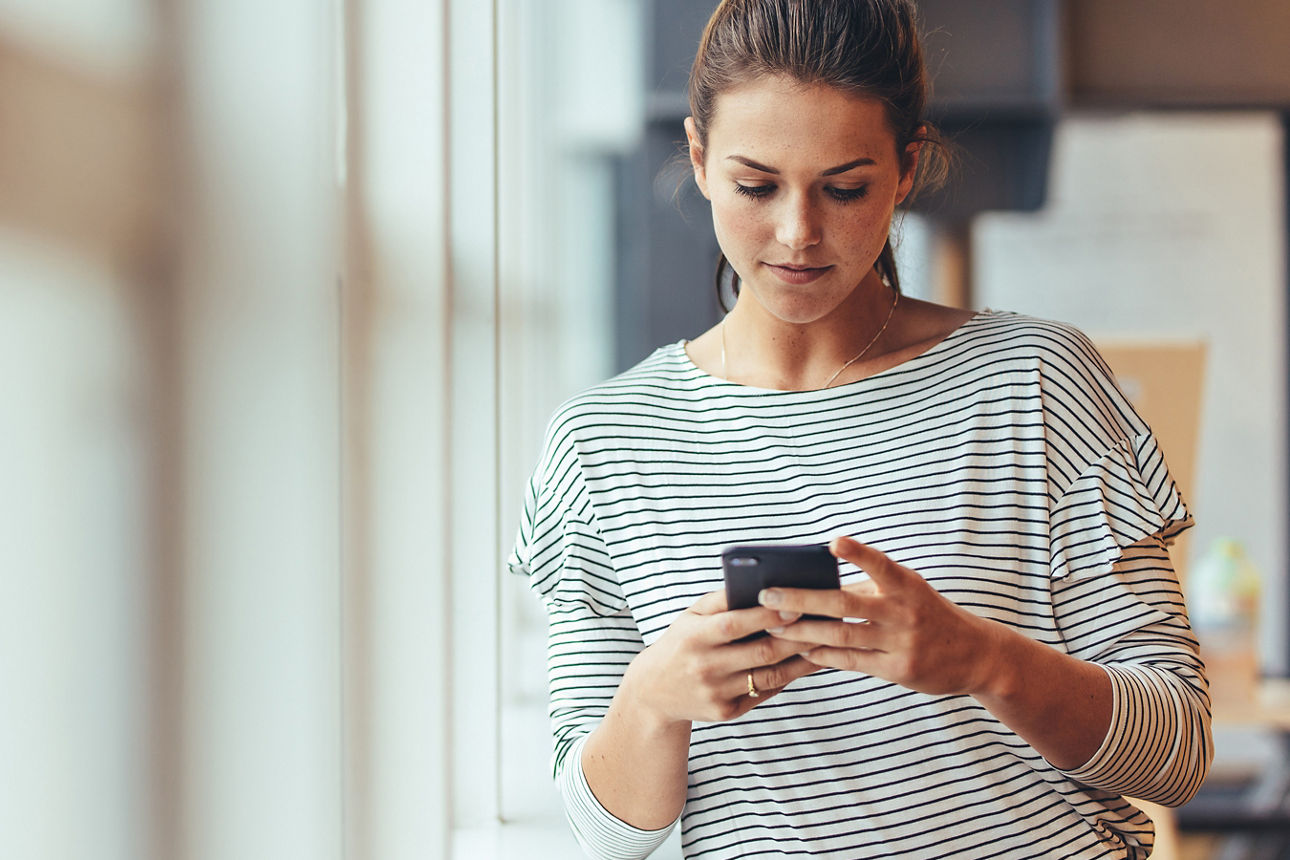 Eine junge Frau mit einem gestreiften Oberteil lehnt sich an der Fensterbank und schaut auf ihr Handy