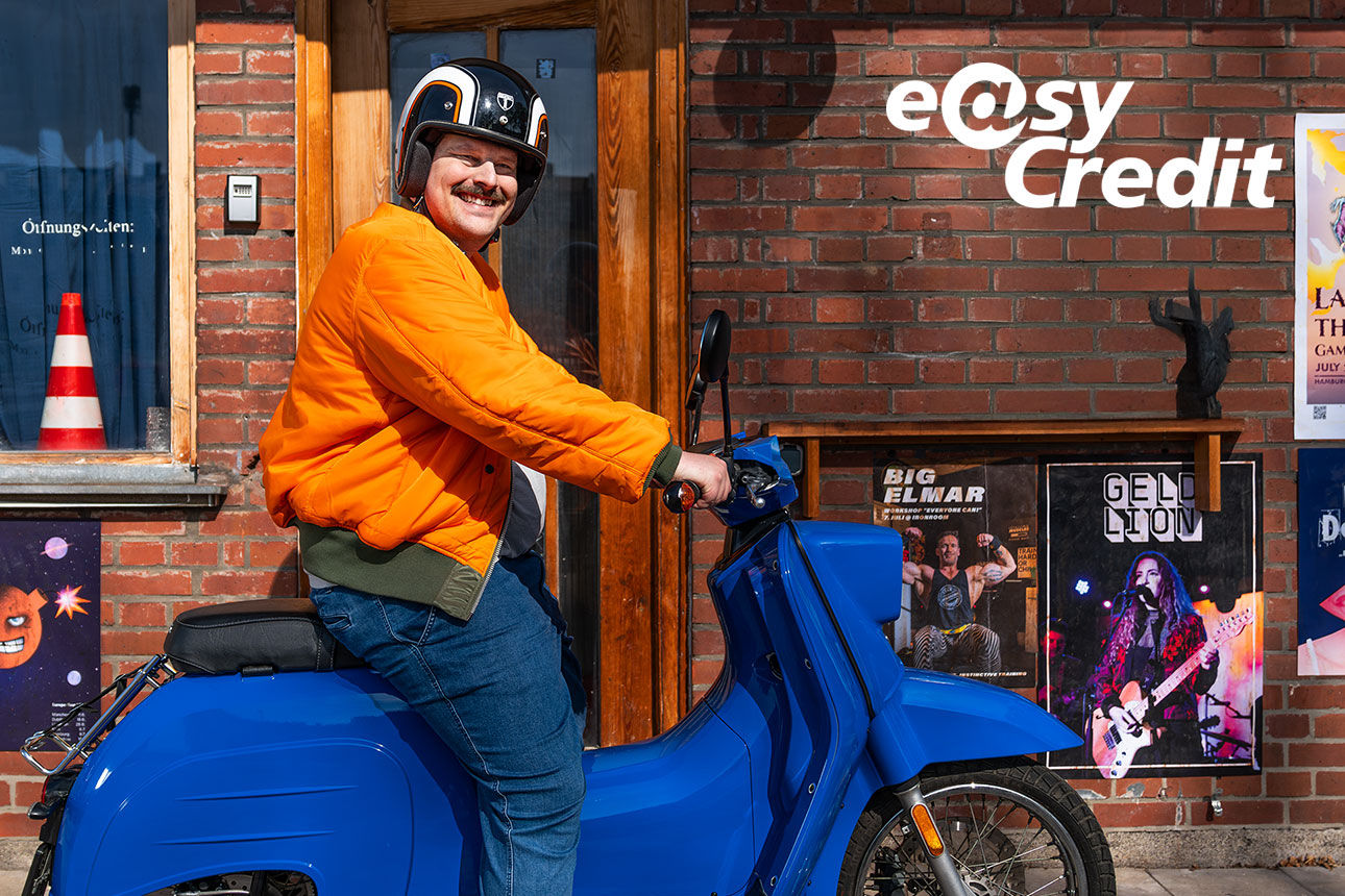 Bild zeigt einen Mann mit oranger Jacke auf einem blauen Roller