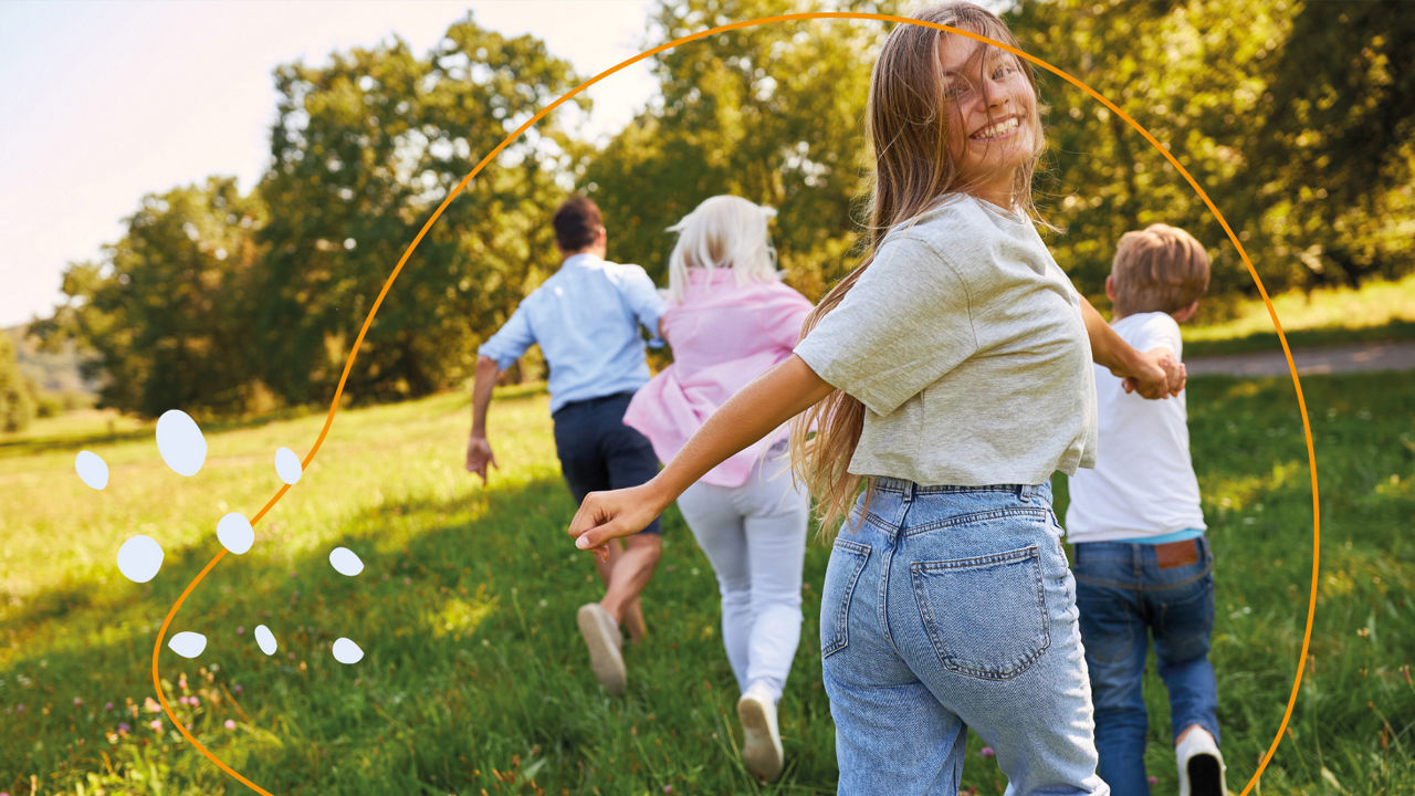 Fröhliches Mädchen guckt in die Kamera und läuft mit anderen Kindern über eine Wiese
