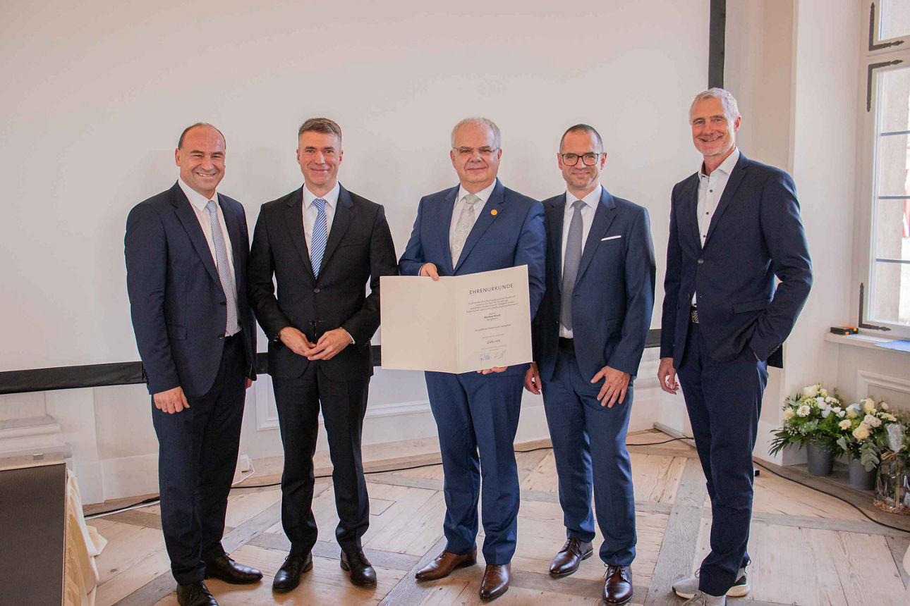 Fünf Personen in Anzügen stehen nebeneinander und lächeln in die Kamera. Eine Person in der Mitte hält eine große Urkunde ode