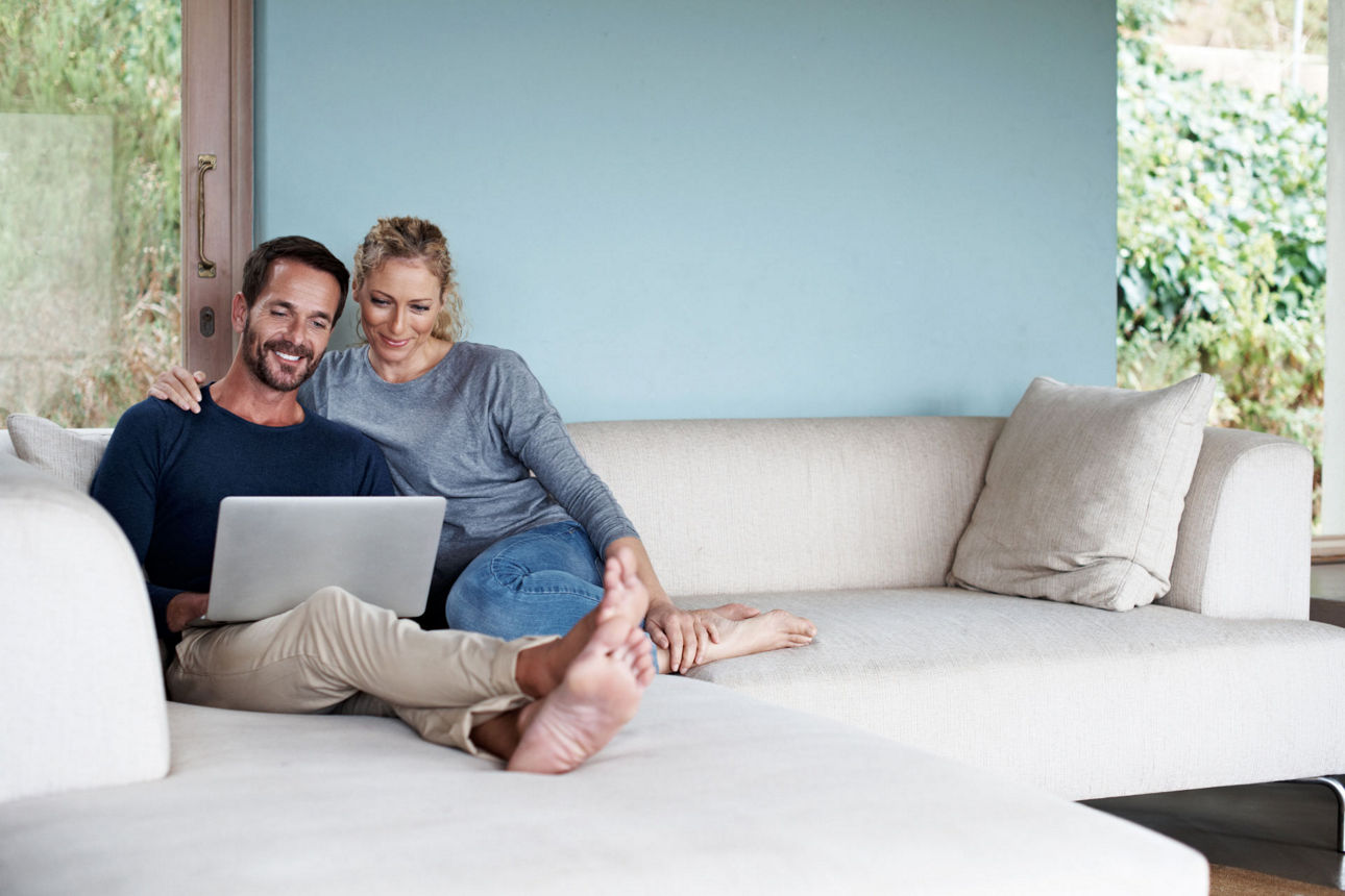 Ein entspanntes Paar sitzt gemeinsam auf einem hellen Sofa in einem modernen Wohnzimmer und blickt lächelnd auf einen Laptop.