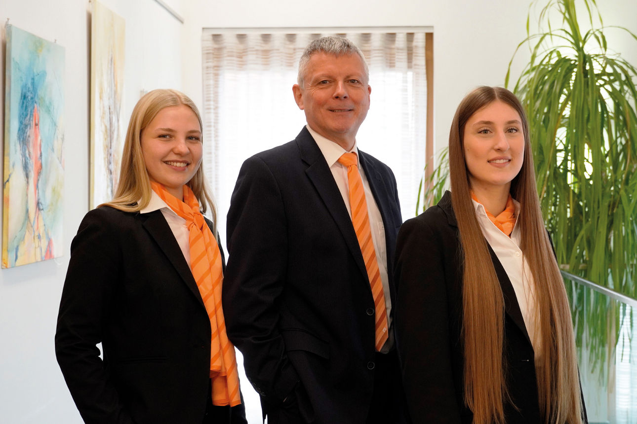 Zwei Frauen und ein Mann in Geschäftskleidung stehen gemeinsam in einem hellen Büroflur.