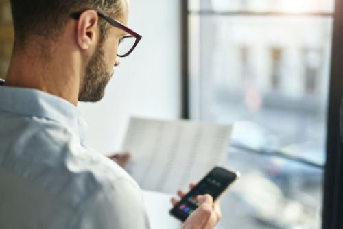 Mann mit Brille hält in der einen Hand Unterlagen und in der anderen ein Smartphone