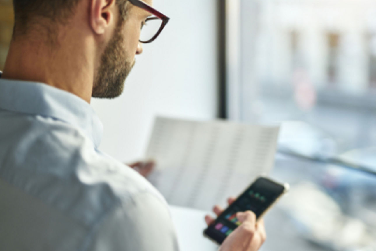 Mann mit Brille hält in der einen Hand Unterlagen und in der anderen ein Smartphone