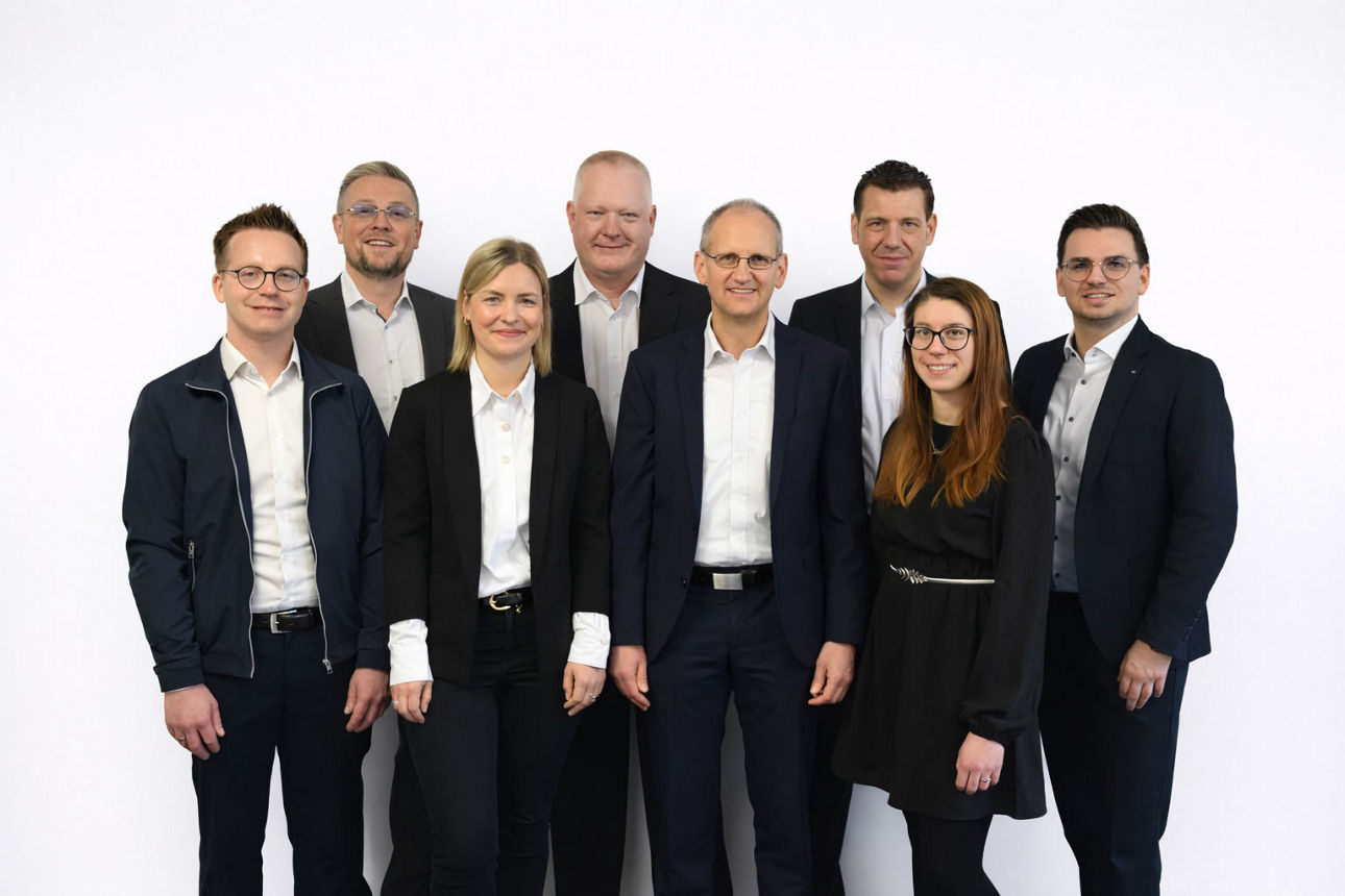 Gruppe von Beraterinnen und Berater in formeller Kleidung steht gemeinsam vor neutralem Hintergrund.