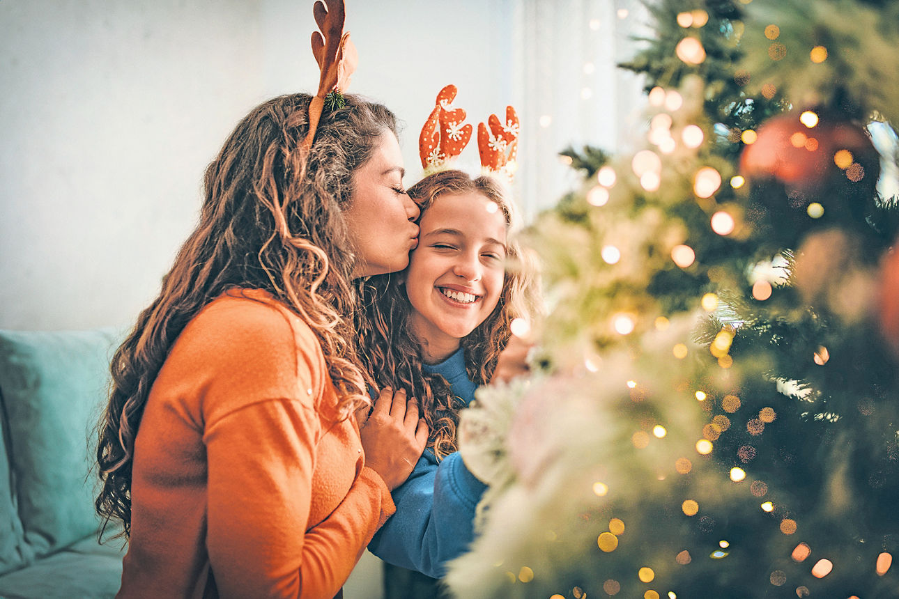 Frau und Kind mit Rentiergeweihen lachen gemeinsam vor einem geschmückten Weihnachtsbaum.