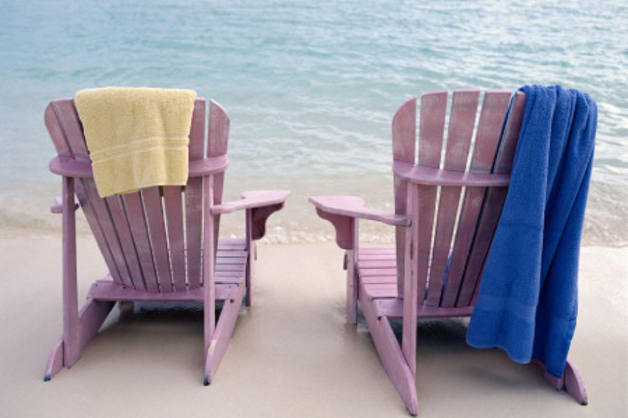 Zwei Sonnenstühle stehen am Strand. Es liegen Handtücher auf den Stühlen. Man sieht das Meer im Hintergrund.