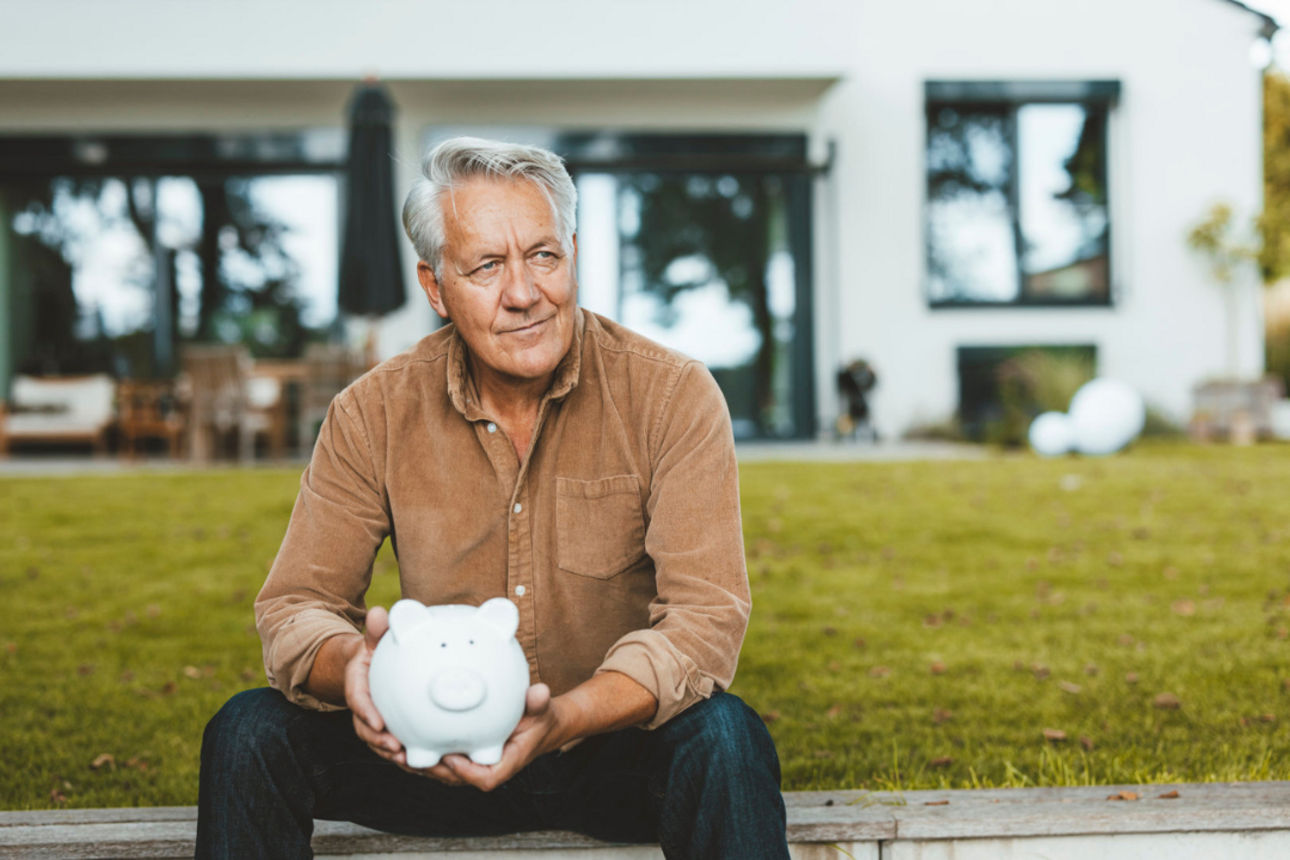 Älterer Herr sitzend vor einem Haus mit einem Sparschwein in der Hand.