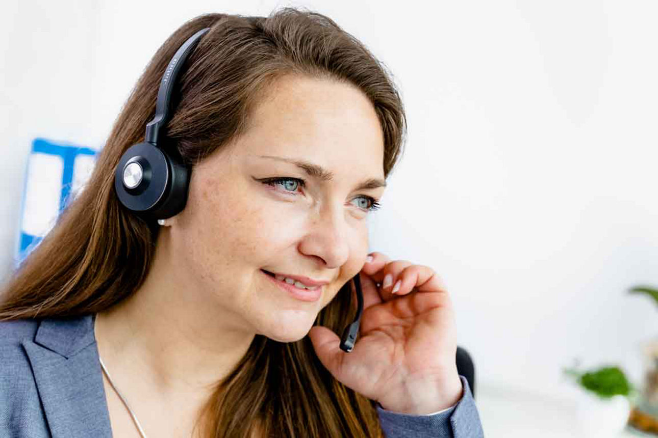 Eine freundliche Frau mit Headset telefoniert konzentriert im hellen Büro und lächelt dabei.