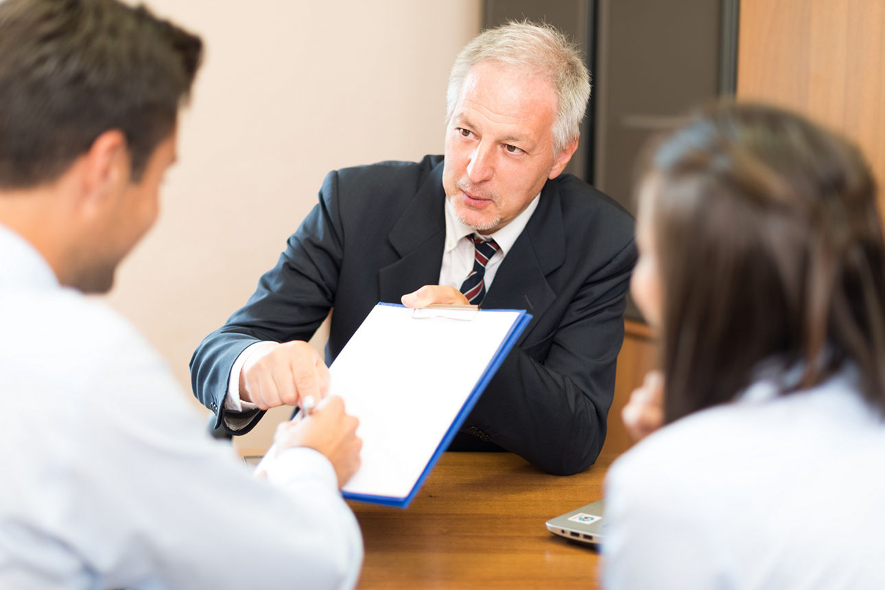 3 Personen sitzen am Tisch, einer unterschreibt Dokument