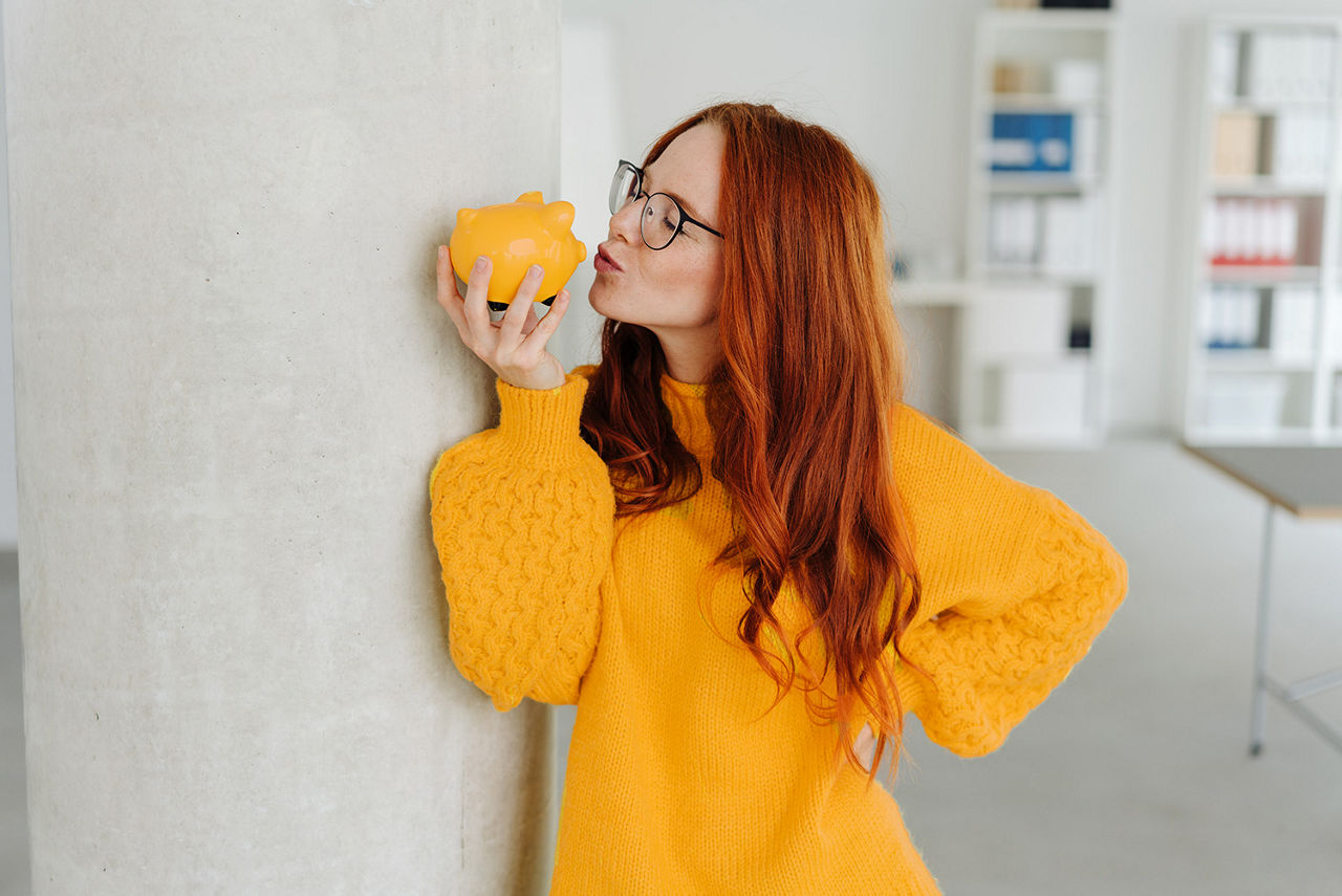 Frau lehnt an der Wand und hält ein Sparschwein in der Hand, das sie küssen will.