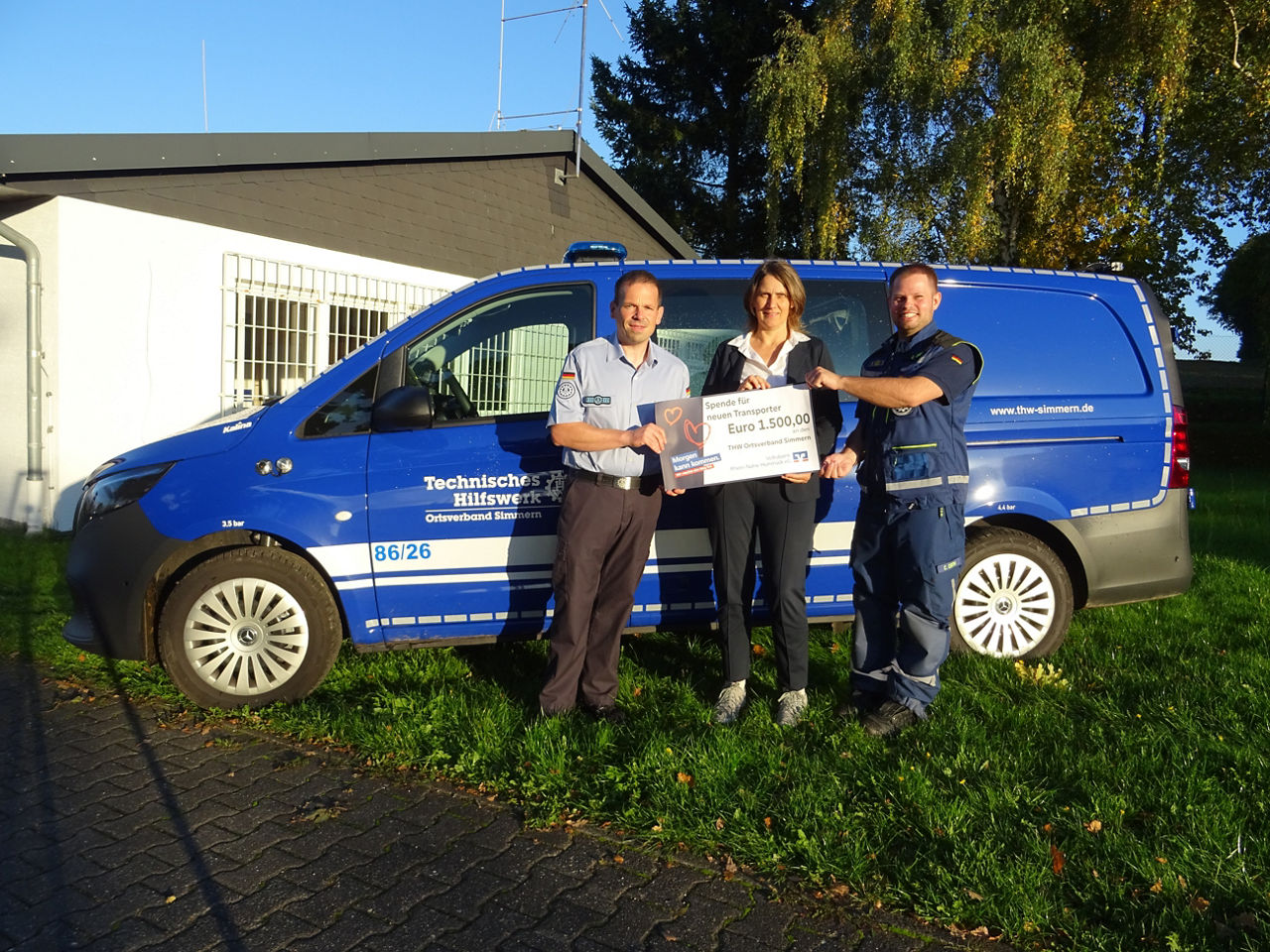 Drei Erwachsene halten den Spendenscheck in der Hand und stehen vor einem blauen Transporter.