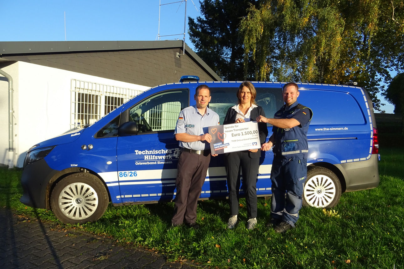 Drei Erwachsene halten den Spendenscheck in der Hand und stehen vor einem blauen Transporter.