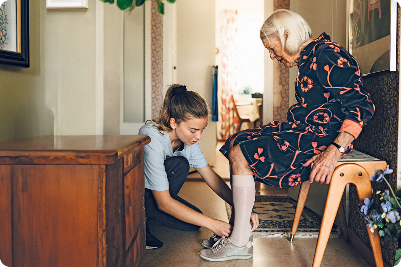 Eine junge Frau kniet vor einer sitzenden älteren Dame und zieht ihr die Schuhe an
