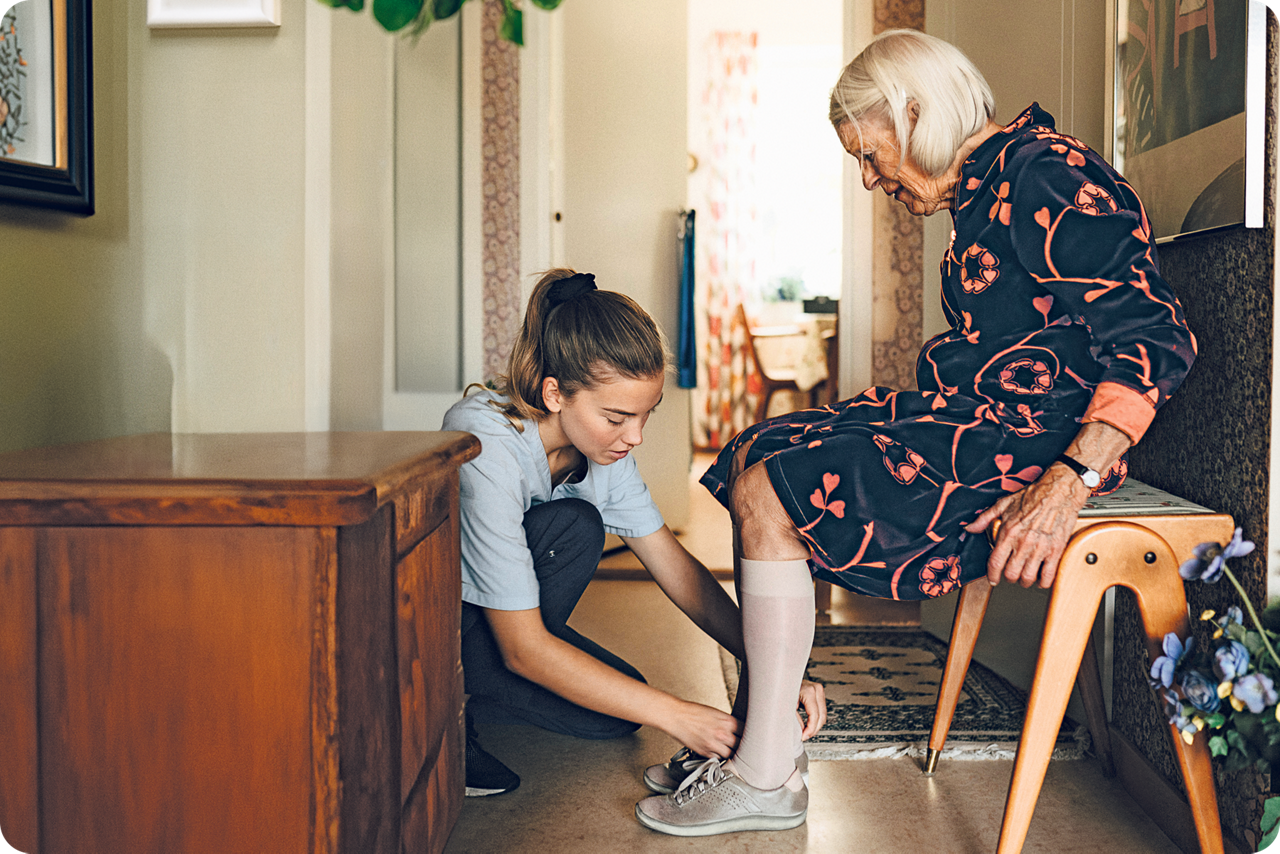 Eine junge Frau kniet vor einer sitzenden älteren Dame und zieht ihr die Schuhe an