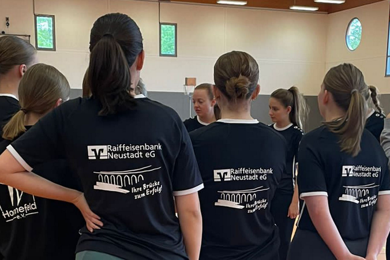 Eine Gruppe von Mädchen mit Zöpfen und Dutt trägt schwarze Raiba-Trikots und steht in einer Turnhalle