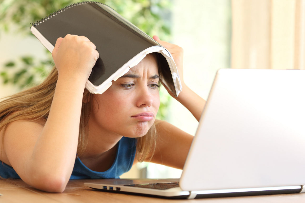 Mädchen schaut genervt in Laptop und hält sich die Hausaufgaben über den Kopf